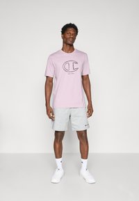 Lila T-Shirt mit schwarzem Logo und Text auf der Brust, kombiniert mit grauen Shorts. Das Modell trägt weiße Sneakers und Knöchel-Socken. Einfaches Design.