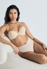 Beiger gepolsterter BH mit dünnen Trägern und passenden hochgeschnittenen Slips. Glatte Textur und nahtloses Design. Das Model liegt auf einer strukturierten weißen Oberfläche.