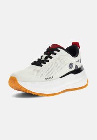 Chaussure de sport blanche avec lacets noirs, tige en mesh texturé, semelle orange et accents imprimés. Présente une languette rouge au talon et le logo "GUESS".