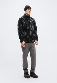 Μαύρο και γκρι φλις μπουφάν με σχέδια, ψηλός γιακάς, φερμουάρ στο μισό, φορεμένο με γκρι παντελόνι και μαύρα παπούτσια πεζοπορίας. Μαύρο πλεκτό σκουφάκι.