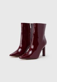 Bottes cheville en cuir verni bordeaux foncé brillant, avec bouts pointus, fermetures éclairs latérales et talons hauts carrés sur fond blanc.