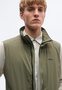 Lekka kamizelka w oliwkowej zieleni z wysokim kołnierzem, zamkiem z przodu i czarnym akcentem. Nałożona na białą koszulę z cienkimi pionowymi paskami.