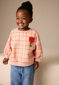 Meruňková mikina s oranžovým mřížkovým vzorem a květinovou výšivkou na levé straně hrudi; doplněná volnými modrými džínami.