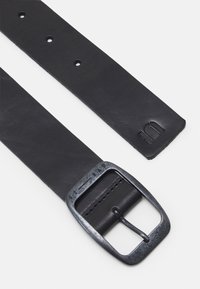 Svart läderbälte med en texturerad yta, utrustat med en metallisk silverfärgad spänne och justeringshål. Varumärke präglat på remmen.