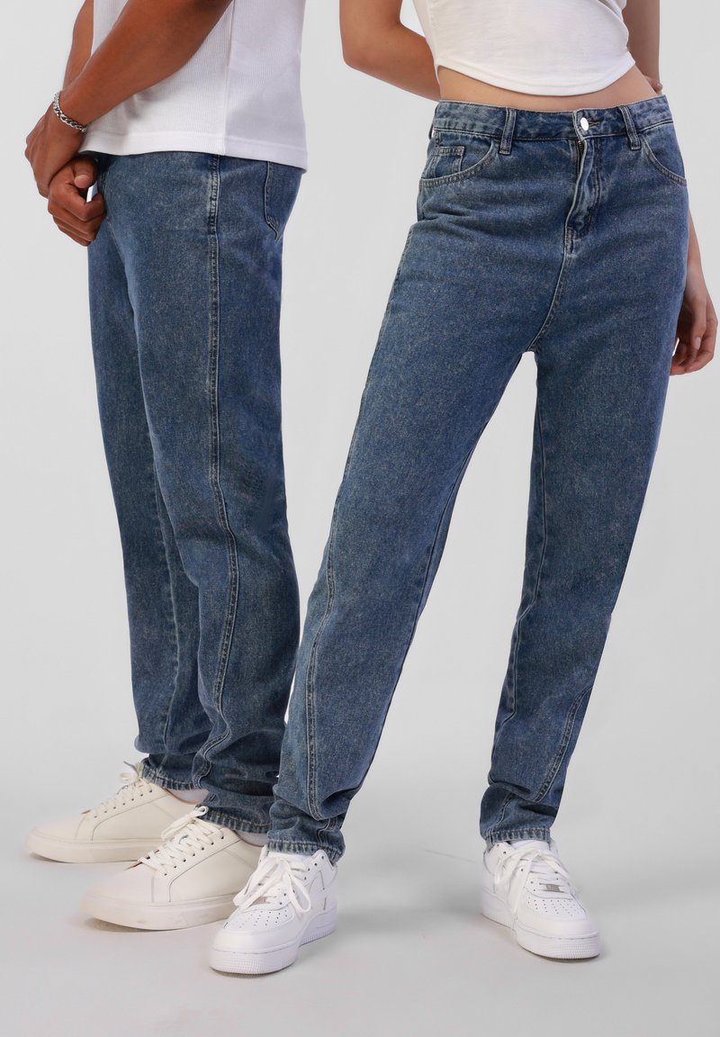 Deux personnes debout côte à côte portant des jeans en denim bleu, des hauts blancs et des baskets blanches sur un fond uni.