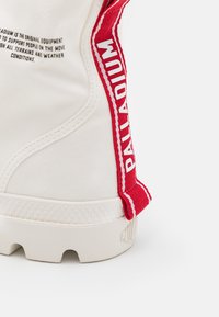 Botte de couleur crème avec une semelle en caoutchouc texturée. Elle possède une sangle d'accent rouge sur le côté avec des lettrages blancs et un talon renforcé.