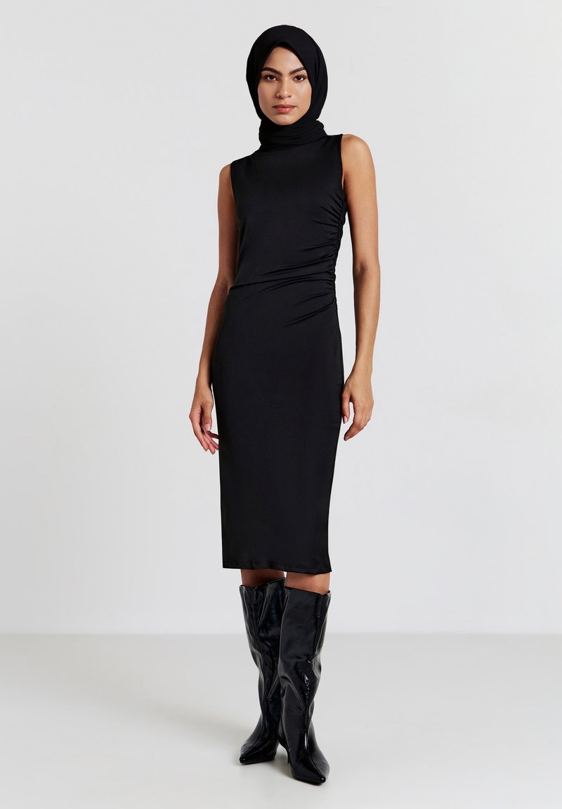 Donna in un vestito midi nero senza maniche, con hijab nero e stivali neri al ginocchio, in piedi contro uno sfondo chiaro e semplice.