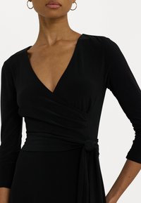 Black wrap dress with long sleeves and a deep V-neck, featuring a knotted detail at the waist and smooth, stretchy fabric.