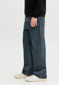 Személy laza sötétkék farmernadrágot, fehér tornacipőt és fekete hosszú ujjú felsőt visel, és sima háttér előtt áll.