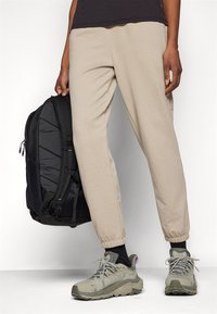 Beige Jogginghosen mit elastischen Bündchen, kombiniert mit grauen Wanderschuhen und einem schwarzen Rucksack mit strukturierten Paneelen und Reißverschluss-Verschlüssen.