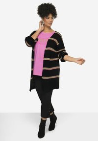 Svart stickad cardigan med flerfärgade ränder, kombinerad med en rosa grafisk t-shirt och svarta capribyxor med snörning vid fållen.