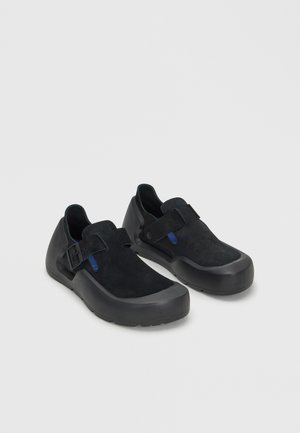Paire de chaussures noires sans lacets avec semelles en caoutchouc et brides réglables, présentées sur un fond gris clair.