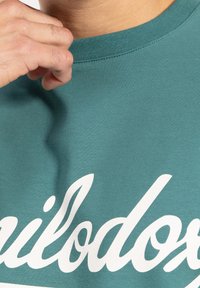 Camiseta de cuello redondo en color turquesa, hecha de un tejido suave, con un gran diseño de texto blanco en la parte delantera. Textura suave con detalle de cuello acanalado.