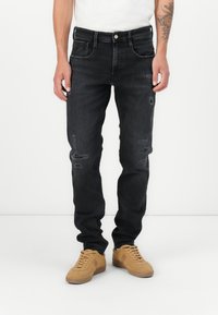 Jeans in denim nero con una vestibilità slim, caratterizzati da dettagli strappati e un classico design a cinque tasche. Indossati con sneakers beige e una camicia bianca.