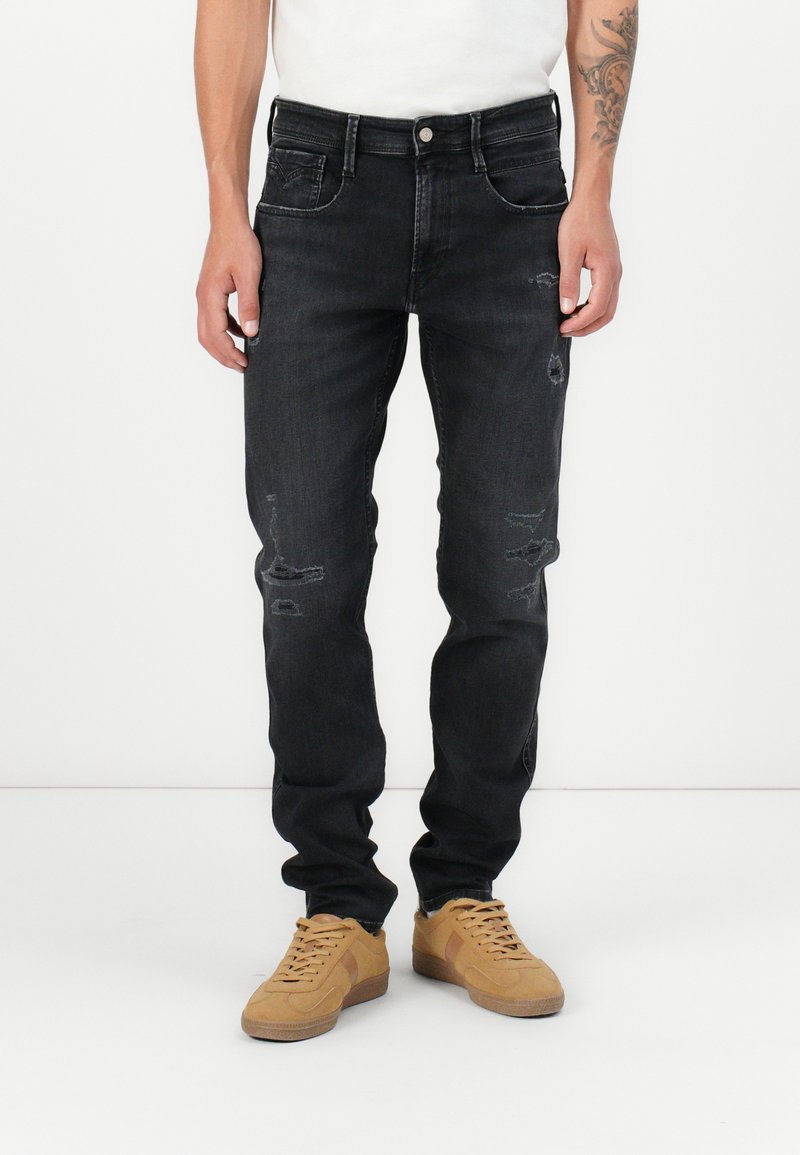 Jeans in denim nero con una vestibilità slim, caratterizzati da dettagli strappati e un classico design a cinque tasche. Indossati con sneakers beige e una camicia bianca.