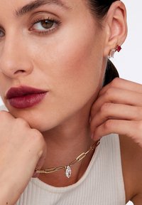 Złota naszyjnik łańcuchowy z wydłużonymi ogniwami i wisiorem w kształcie marquise z kryształu. Modelka nosi małe kolczyki wkrętki w różnych kolorach.