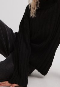 Pull à col roulé noir en maille côtelée avec manches longues, coupe décontractée et tissu texturé, associé à un pantalon gris foncé.