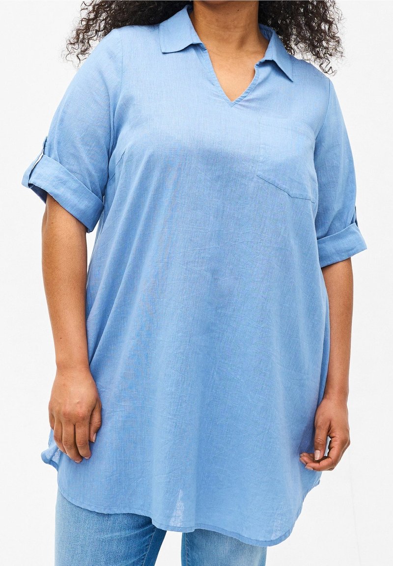 Femme portant une tunique bleu clair ample avec manches retroussées et col par-dessus un jean bleu sur fond blanc.