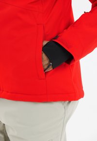 Main dans la poche d'une veste rouge vif avec poignet noir, pantalon beige visible en dessous, gros plan sur le torse latéral et le bras.