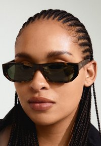 Lunettes de soleil en écaille de tortue avec des verres foncés, au design carré et à monture épaisse. Présentent une branding subtile sur les branches. Texture lisse.