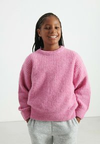 Ružový, štruktúrovaný pletený sveter s okrúhlym výstrihom a klesajúcimi ramenami, kombinovaný so šedými teplákmi. Materiál vyzerá mäkko a teplo.
