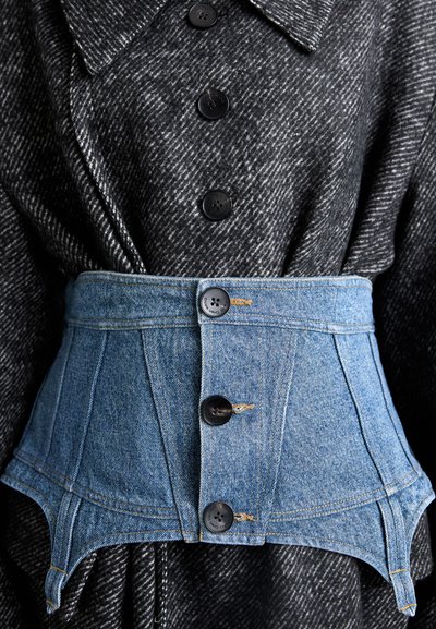 Cinturón corsé de denim con cuatro botones negros, llevado sobre una tela de tejido de pata de gallo en blanco y negro, con cuello y mangas largas.