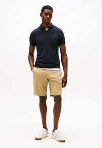 Tamsiai mėlynos spalvos polo marškinėliai, chaki spalvos šortai ir balti sportbačiai. Lygus medvilninis audinys, klasikinis kirpimas ir subtili logotipo detalė ant polo marškinėlių.