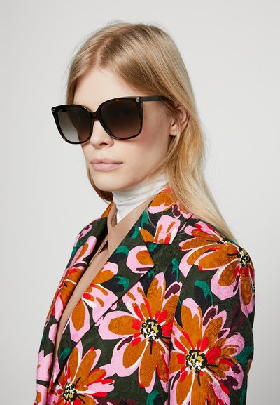 Ochelari de soare oversize negri cu lentile în degrade, asortați cu un sacou îndrăzneț cu un model floral, având flori mari roz și portocalii pe un fundal închis.