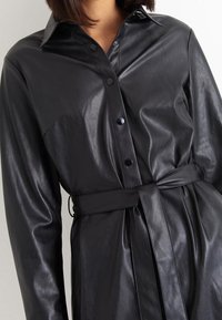 Abito in pelle nera con chiusura a bottoni sul davanti, maniche lunghe e vita cinturata. Caratterizzato da un colletto classico e una texture liscia.