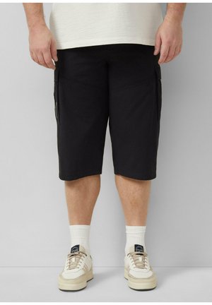 Man draagt zwarte cargoshorts, witte sportsokken en beige sneakers, staand tegen een effen lichte achtergrond.