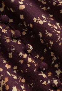 Tissu violet foncé avec un petit motif floral beige et des boutons recouverts de tissu cousus en ligne verticale créant des fronces.