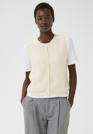 Gebreide mouwloze sweatervest in zacht crème, met een ronde halslijn en knoopsluiting. Draag het over een wit T-shirt met korte mouwen en grijs gestreepte broek.