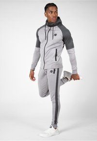 Siva i tamno siva zip-up dukserica s elastičnim manžetama, uparena s odgovarajućim trenirkama s bočnim prugama i logotipom. Nosena s bijelim tenisicama.