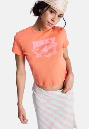 Młoda kobieta w różowym kwiatowym chustce na głowie, koralowym krótkim t-shircie Roxy i pastelowej spódnicy w paski, pozująca z ręką na biodrze.