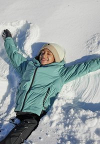 Ein Kind in einer mintgrünen Daunenjacke liegt im frischen, weißen Schnee, trägt eine hellbeige Mütze und schwarze Handschuhe, die Arme weit ausgestreckt.