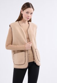 Gilet en polaire beige avec des bordures en imprimé léopard, devant ouvert, deux poches plaquées et sans manches, porté sur un haut à manches longues beige clair.