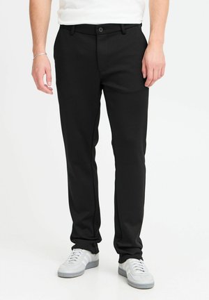 Svarte bukser med rett ben og med en glatt stofffinish, med knapplås i livet og sidelommer. Bært med hvite sneakers.