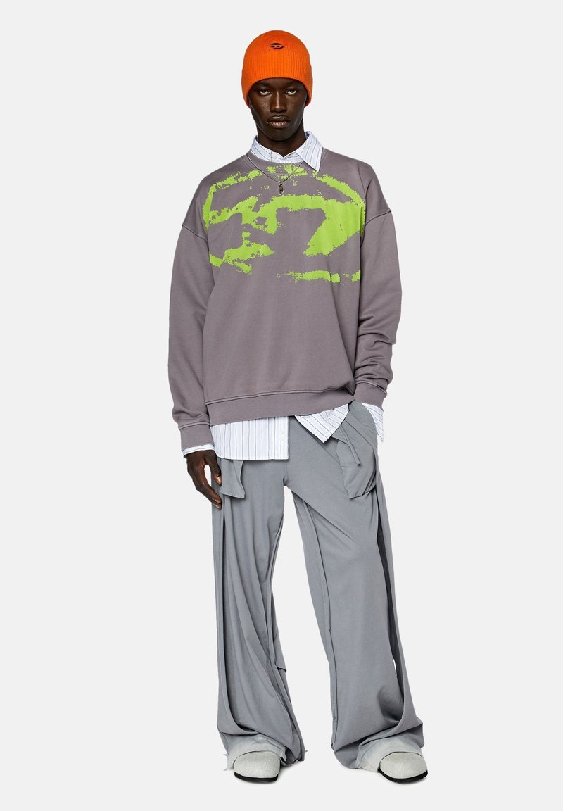 Sweatshirt gris surdimensionné avec un motif abstrait vert, superposé à une chemise rayée blanche ; associé à un pantalon gris ample et un bonnet orange.