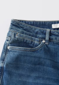 Jean en denim bleu à taille haute, avec des surpiqûres cuivrées et une poche frontale. Présente un bouton et une étiquette au niveau de la taille.