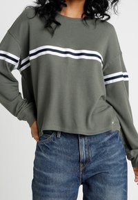 Olivgrön ribbad tröja med vita och marinblå horisontella ränder över bröstet. Har långa ärmar och en avslappnad passform. Matchas med jeans.