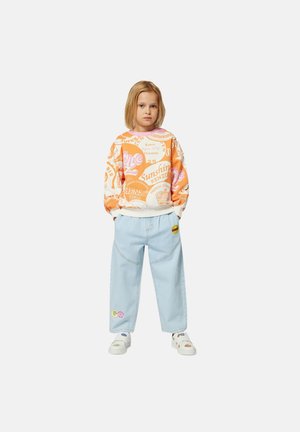 Helles oranges Sweatshirt mit verschiedenen Logos, kombiniert mit hellblauen Jeans, die bunte Patches aufweisen, und weißen Slip-On-Schuhen.