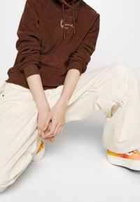 Sudadera marrón con capucha, acabado texturizado y detalle bordado, combinada con pantalones blancos y sueltos y zapatillas multicolores.