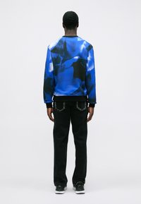Hombre de pie con la espalda hacia la cámara, vistiendo una sudadera con un patrón azul y negro, jeans negros y zapatillas negras con suelas blancas.