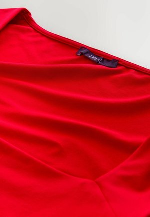 Roter Stoff mit glatter Textur, der einen tiefen V-Ausschnitt und ein gefaltetes Design aufweist. Ein dunkles Etikett mit der Aufschrift "next" ist in der Nähe des Ausschnitts sichtbar.