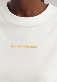 Hvid sweatshirt med ribbet krave, og "MarcOPoloDenim" broderet i orange på brystet. Blød stoftekstur.