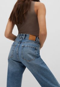 Mujer vistiendo jeans de tiro alto azules y una blusa sin mangas marrón, de pie con una mano en la cadera frente a un fondo sencillo.