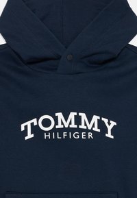 Μπλούζα φούτερ σε ναυτικό μπλε χρώμα με μαλακή υφή, που διαθέτει μεγάλο λευκό λογότυπο "TOMMY HILFIGER". Η κουκούλα είναι σχεδιασμένη με κλείσιμο βελόνας μπροστά.