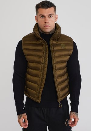 Uomo con tatuaggi che indossa un gilet imbottito verde oliva sopra un maglione nero e pantaloni neri, in piedi davanti a uno sfondo neutro.