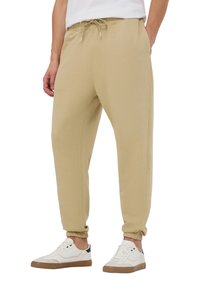 Pantaloni sportivi beige con vita elastica e cordoncino, dotati di caviglie a costo e tasche laterali, abbinati a scarpe da tennis bianche.