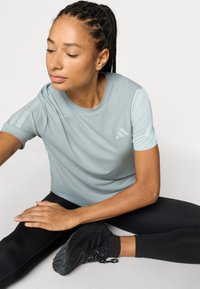 Jasnoniebieska sportowa koszulka z krótkimi rękawami, z trzema białymi paskami na ramionach i małym logo na piersi. Czarne legginsy i sneakersy.